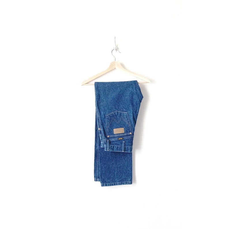 May include: A pair of blue denim jeans hanging on a wooden hanger against a white background. The jeans have a brown leather patch on the back pocket with the word 'Wrangler' printed on it.