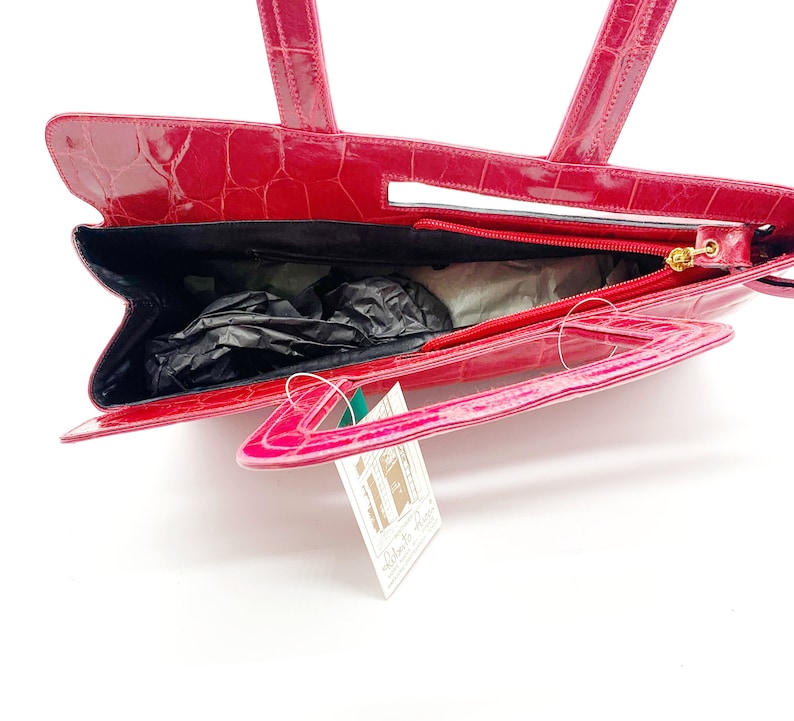 May include: A pink crocodile embossed leather handbag with a top handle and a detachable shoulder strap. The bag has a zipper closure and a gold-toned hardware.