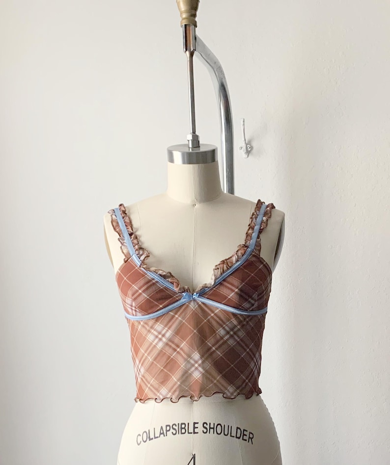 May include: A brown plaid camisole top with a light blue trim and straps. The top has a ruffled edge and a small bow detail at the center. The garment is displayed on a mannequin.