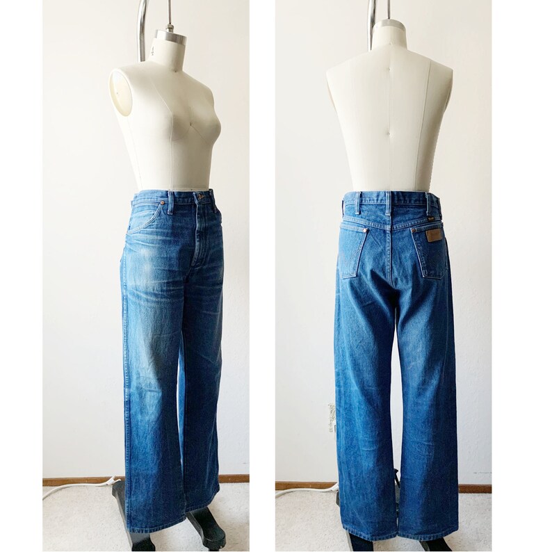 May include: A pair of blue denim jeans with a wide leg and a high waist. The jeans have a brown leather patch on the back pocket with the word "Wrangler" printed on it.