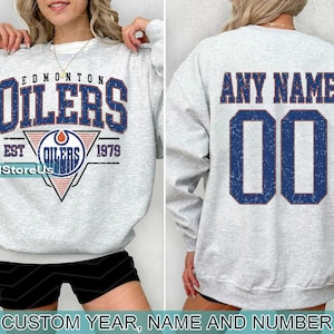 May include: A gray sweatshirt with the text "Edmonton Oilers Est. 1979" and a logo. The back of the sweatshirt has the text "Any Name" and a number.