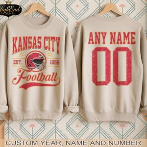 Puede incluir: Sudadera beige con un gr&aacute;fico de "Kansas City Football" en la parte delantera, con un casco de f&uacute;tbol americano rojo. La parte trasera muestra "ANY NAME" y el n&uacute;mero "00" en rojo. Opciones personalizadas de a&ntilde;o, nombre y n&uacute;mero.