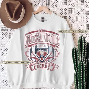 K&ouml;nnte beinhalten: Wei&szlig;es Sweatshirt mit roter und blauer Grafik mit der Aufschrift "2023 MONTREAL VICTOIRE HOCKEY". Das Design beinhaltet ein ovales Emblem mit einem Schild und einem Banner. Ein brauner Hut h&auml;ngt links.