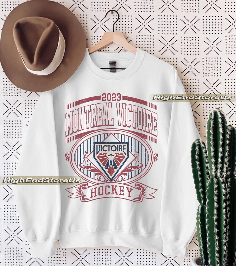 K&ouml;nnte beinhalten: Wei&szlig;es Sweatshirt mit roter und blauer Grafik mit der Aufschrift "2023 MONTREAL VICTOIRE HOCKEY". Das Design beinhaltet ein ovales Emblem mit einem Schild und einem Banner. Ein brauner Hut h&auml;ngt links.