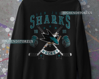 Puede incluir: Sudadera negra con un logotipo teal de los San Jose Sharks. El logotipo presenta un tiburón con palos de hockey cruzados y las palabras "San Jose Sharks" y "Hockey" en blanco.