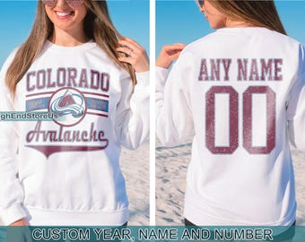 May include: White sweatshirt with a burgundy Colorado Avalanche logo on the front and a burgundy "ANY NAME" and number 00 on the back.  The logo includes the text "Colorado Avalanche" and "EST. 1972".