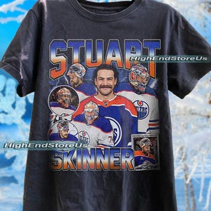 May include: Dark gray t-shirt featuring a collage of images of Stuart Skinner, a hockey player, with the name "STUART SKINNER" in orange and blue lettering. The shirt also has the text "HighEndStoreUs".