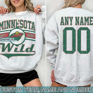Pode incluir: Uma camisa de moletom cinza com o texto "MINNESOTA EST. 2000 Wild" e um logotipo verde e branco. A parte de trás da camisa de moletom tem o texto "ANY NAME" e o número "00" em verde.