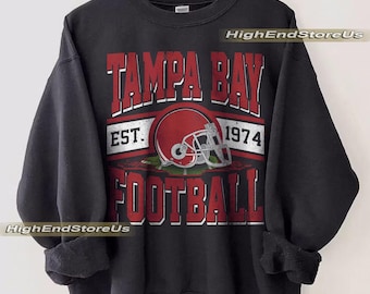 Puede incluir: Sudadera negra con un gráfico rojo y blanco. El gráfico dice "TAMPA BAY FOOTBALL EST. 1974" con un casco de fútbol americano rojo en el centro. La sudadera está colgada en una percha de madera.