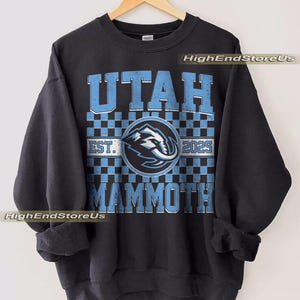 Puede incluir: Sudadera negra con la palabra "UTAH" en letras de bloque azul claro sobre un patrón de cuadros. Debajo del patrón está la palabra "MAMMOTH" en azul claro. Un emblema circular con un gráfico de mamut está en el centro. También es visible el texto "EST. 2025".