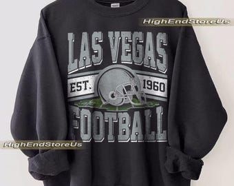 Può includere: Felpa nera con un design grafico invecchiato. Il design presenta le parole "LAS VEGAS FOOTBALL" con un casco da football al centro. È incluso anche il testo "EST. 1960". La felpa è appesa a una gruccia di legno.