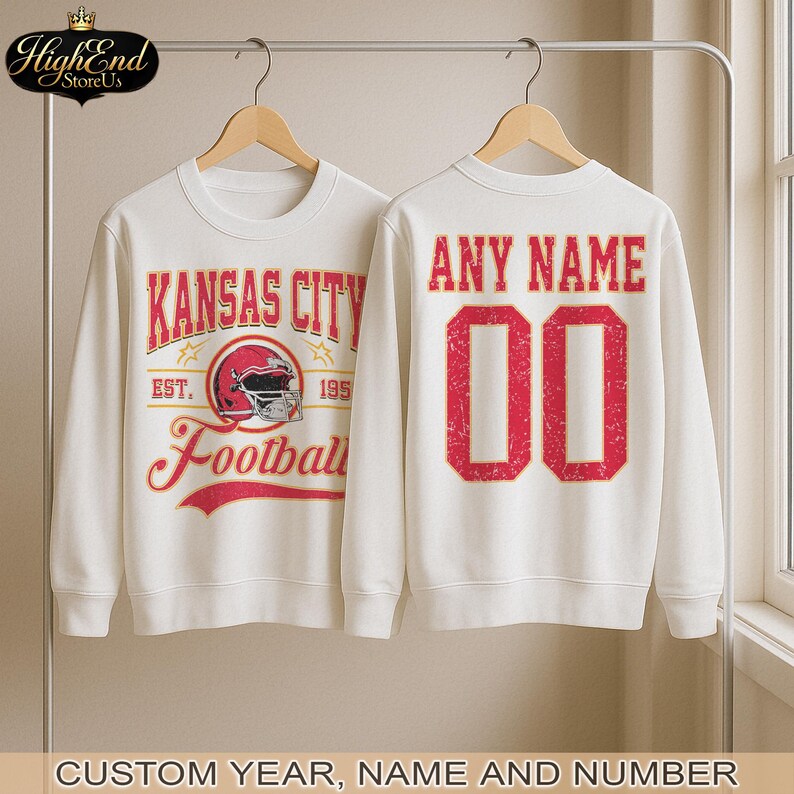 Puede incluir: Dos sudaderas blancas en perchas. Una presenta "Kansas City Football" en rojo y blanco, con un gr&aacute;fico de casco de f&uacute;tbol. La otra tiene "ANY NAME" y el n&uacute;mero "00" en rojo. El texto inferior dice "CUSTOM YEAR, NAME AND NUMBER."