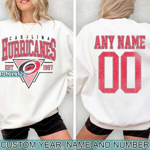 May include: White sweatshirt with a red and black Carolina Hurricanes logo and the text "Carolina Hurricanes Est 1997". The back of the sweatshirt has the text "Any Name" and a red number "00".