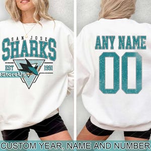 May include: White sweatshirt with a teal blue San Jose Sharks logo and the text "San Jose Sharks Est 1991". The back of the sweatshirt has the text "Any Name 00" in teal blue.