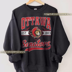 Puede incluir: Sudadera negra de los Senators de Ottawa con el logotipo y el texto del equipo en rojo y blanco. El diseño incluye el nombre del equipo, el logotipo y el año de fundación, 1883. La sudadera está colgada en una percha de madera.