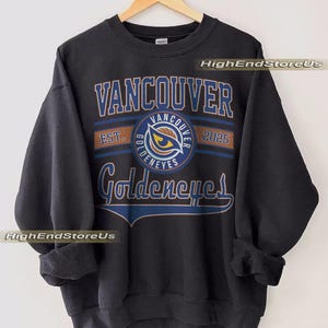May include: Black crew neck sweatshirt featuring the text "Vancouver Goldeneyes" in blue and gold. The design includes a circular emblem with an eye graphic and the text "Est. 2025". The sweatshirt is displayed on a wooden hanger.
