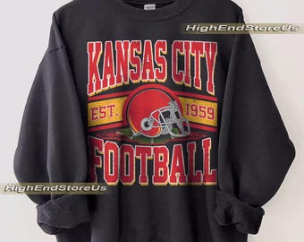 Peut inclure: Sweat-shirt noir avec un motif graphique portant l'inscription "KANSAS CITY FOOTBALL" en rouge, soulignée de blanc. Un casque de football rouge est centré dans le motif, avec l'année "1959" imprimée au-dessus. Le sweat-shirt est suspendu à un cintre en bois.