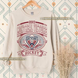 K&ouml;nnte beinhalten: Cremefarbenes Sweatshirt mit einem verblassten rot-blauen Grafikdesign. Der Aufdruck lautet "2023 MONTREAL VICTOIRE HOCKEY" im Vintage-Stil. Das Design umfasst ein Wappen und B&auml;nder, ideal f&uuml;r Hockeyfans.