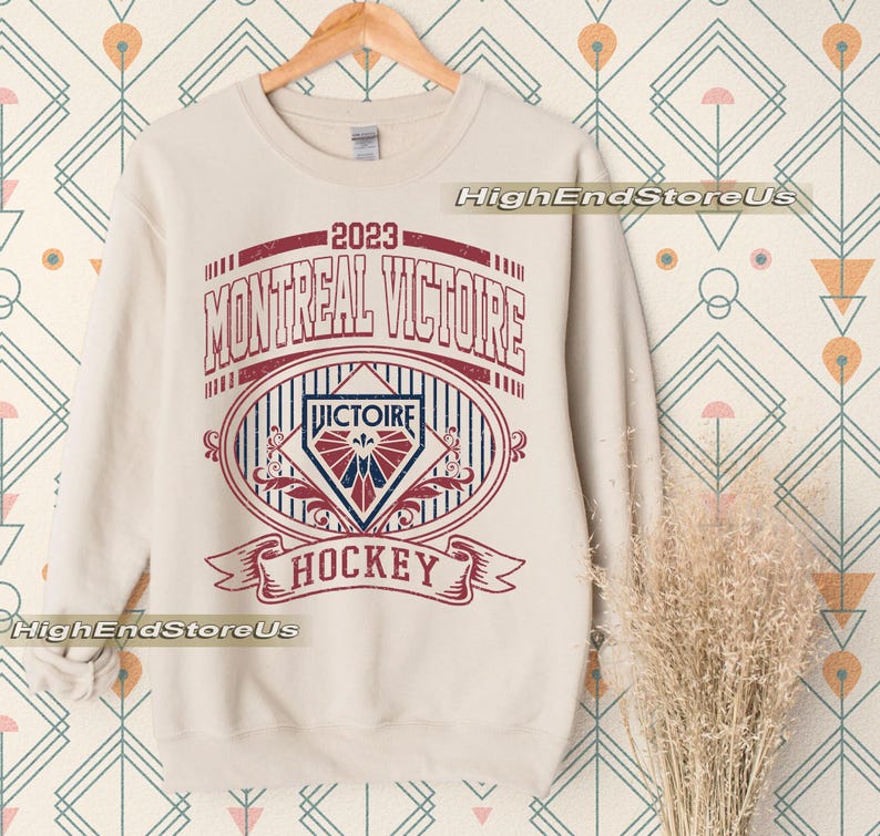 K&ouml;nnte beinhalten: Cremefarbenes Sweatshirt mit einem verblassten rot-blauen Grafikdesign. Der Aufdruck lautet "2023 MONTREAL VICTOIRE HOCKEY" im Vintage-Stil. Das Design umfasst ein Wappen und B&auml;nder, ideal f&uuml;r Hockeyfans.