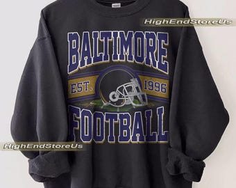 Puede incluir: Sudadera negra con "BALTIMORE FOOTBALL" en letras blancas y azules. Un gráfico de casco de fútbol está centrado debajo del texto, con "EST. 1996" arriba. La sudadera está colgada en una percha de madera.