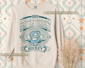 May include: Cream-colored sweatshirt with a teal and white graphic. The graphic reads "2025 SEATTLE TORRENT HOCKEY" in a vintage style. The design includes an oval with the letter "S" and the word "TORRENT".