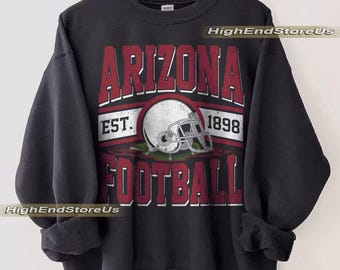 Op de afbeelding: Zwart sweatshirt met een bordeauxrode en witte afbeelding. De afbeelding leest "ARIZONA FOOTBALL EST. 1898" met een voetbalhelm in het midden. Het sweatshirt hangt aan een houten hanger.