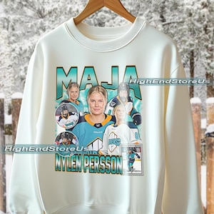 Op de afbeelding: Witte sweatshirt met een turquoise en zwarte afbeelding van een hockeyspeelster genaamd Maja Nylen Persson. Het ontwerp bevat meerdere afbeeldingen van de speler en de tekst "MAJA" en "NYLEN PERSSON". De sweatshirt heeft een ronde hals en lange mouwen.