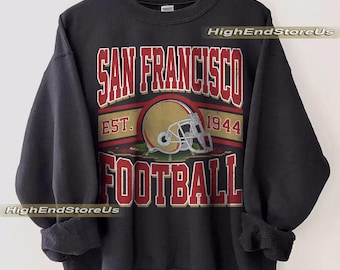 Puede incluir: Sudadera negra con un gráfico desgastado de un casco de fútbol y las palabras "SAN FRANCISCO FOOTBALL" en rojo y blanco. También se incluye el texto "EST. 1944". La sudadera está colgada en una percha de madera.