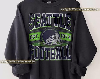 May include: Black sweatshirt featuring the words "SEATTLE FOOTBALL" in white and green block letters. The design includes a football helmet and the text "EST. 1974". The sweatshirt is hanging on a wooden hanger.