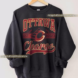 Puede incluir: Sudadera negra con el texto "OTTAWA Charge" en letras rojas y doradas. El diseño incluye una "C" estilizada dentro de un rombo, con "EST. 2023" debajo. La sudadera está colgada en una percha de madera.