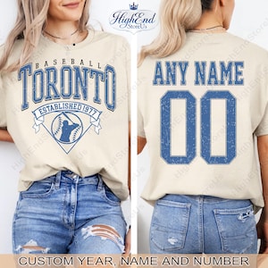Puede incluir: Una camiseta de béisbol beige con la palabra "TORONTO" en azul, junto con "ESTABLISHED 1977" y un gráfico de béisbol. La parte trasera de la camiseta tiene "ANY NAME" y el número "00" en azul. El texto "CUSTOM YEAR, NAME AND NUMBER" está en la parte inferior.