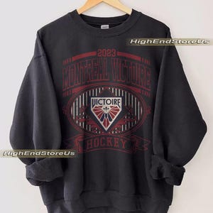 K&ouml;nnte beinhalten: Schwarzes Sweatshirt mit roter und wei&szlig;er Grafik. Die Grafik zeigt den Text "2023 MONTREAL VICTOIRE HOCKEY" und ein rautenf&ouml;rmiges Emblem. Das Sweatshirt h&auml;ngt an einem Holzkleiderb&uuml;gel.