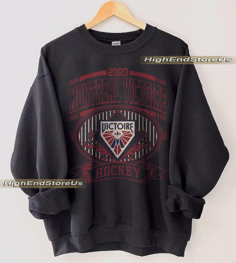 K&ouml;nnte beinhalten: Schwarzes Sweatshirt mit roter und wei&szlig;er Grafik. Die Grafik zeigt den Text "2023 MONTREAL VICTOIRE HOCKEY" und ein rautenf&ouml;rmiges Emblem. Das Sweatshirt h&auml;ngt an einem Holzkleiderb&uuml;gel.