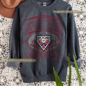 K&ouml;nnte beinhalten: Dunkelgraues Sweatshirt mit roter und wei&szlig;er Grafik. Die Grafik enth&auml;lt den Text "MONTREAL VICTOIRE HOCKEY" und ein Schildemblem. Das Sweatshirt wird auf einem Holzkleiderb&uuml;gel pr&auml;sentiert.