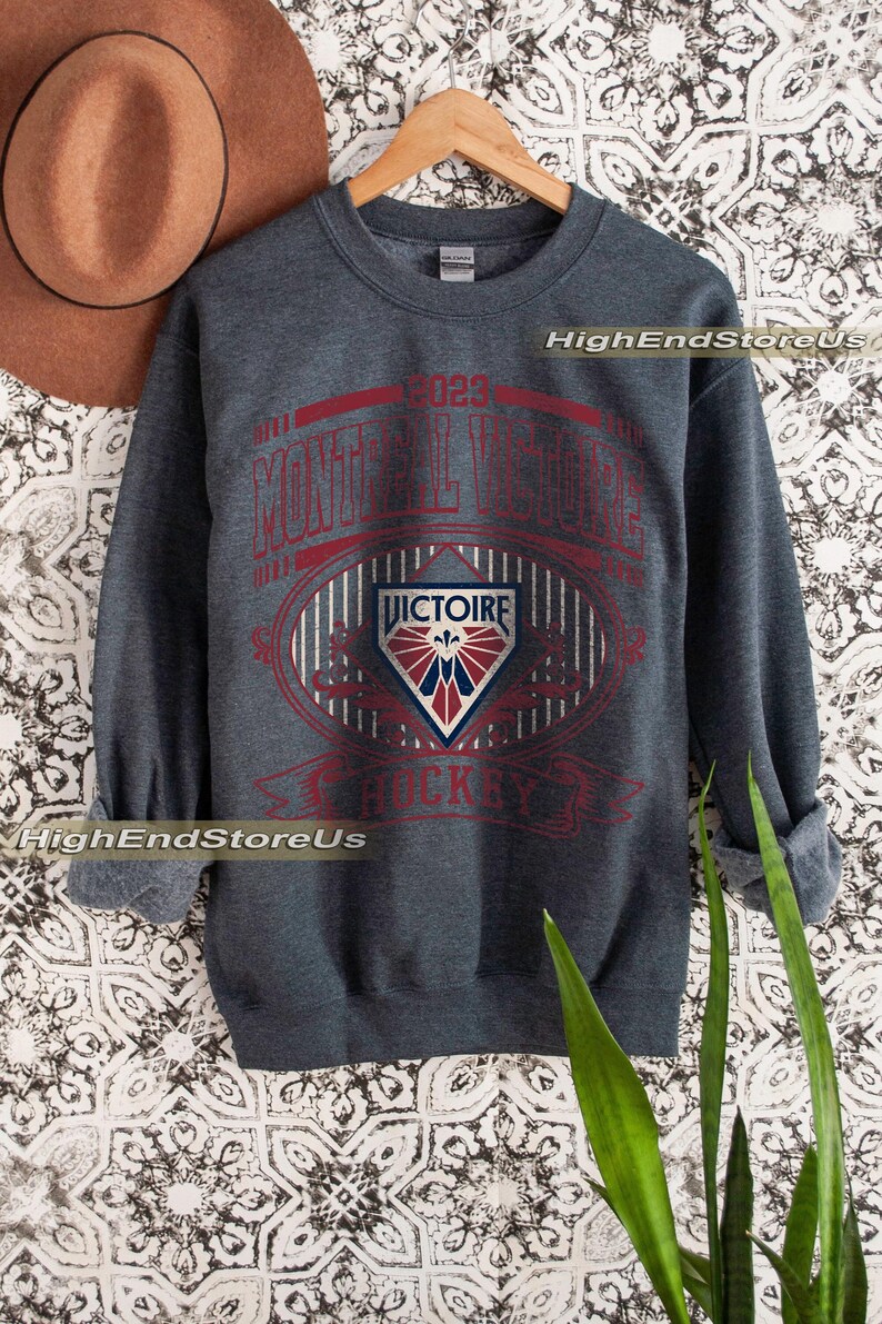 K&ouml;nnte beinhalten: Dunkelgraues Sweatshirt mit roter und wei&szlig;er Grafik. Die Grafik enth&auml;lt den Text "MONTREAL VICTOIRE HOCKEY" und ein Schildemblem. Das Sweatshirt wird auf einem Holzkleiderb&uuml;gel pr&auml;sentiert.