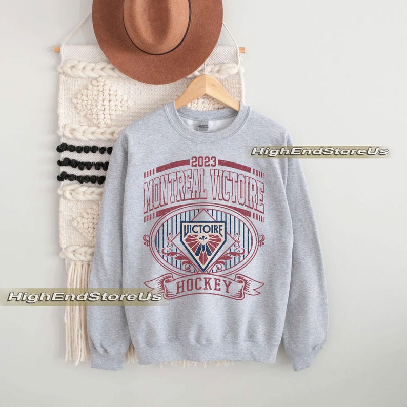K&ouml;nnte beinhalten: Ein meliertes graues Sweatshirt mit dem Text "2023 MONTREAL VICTOIRE HOCKEY" in Rot und Wei&szlig;. Das Design beinhaltet ein ovales Emblem mit einem stilisierten Logo und dem Wort "VICTOIRE". Das Sweatshirt h&auml;ngt an einem Holzkleiderb&uuml;gel.