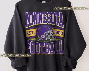 May include: Black sweatshirt with a purple and gold graphic. The graphic reads "MINNESOTA FOOTBALL" with a football helmet and the year "1960". The sweatshirt is hanging on a wooden hanger.