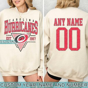 May include: A cream-coloured sweatshirt with a red and black Carolina Hurricanes logo on the front and the text "Carolina Hurricanes Est 1997". The back of the sweatshirt has the text "Any Name 00" in red.
