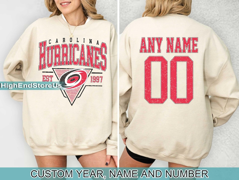 May include: A cream-coloured sweatshirt with a red and black Carolina Hurricanes logo on the front and the text "Carolina Hurricanes Est 1997". The back of the sweatshirt has the text "Any Name 00" in red.