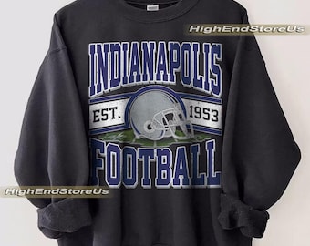 May include: Black sweatshirt with a graphic design featuring the text "INDIANAPOLIS FOOTBALL" in white and blue, with a football helmet graphic. The text "EST. 1953" is also included. The sweatshirt is hanging on a wooden hanger.