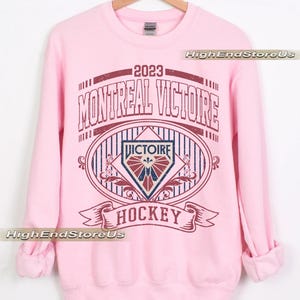 K&ouml;nnte beinhalten: Ein hellrosa Sweatshirt mit einer weinroten und marineblauen Grafik. Die Grafik lautet "2023 MONTREAL VICTOIRE HOCKEY" im Vintage-Stil. Das Sweatshirt ist aus weichem Material und hat lange &Auml;rmel.