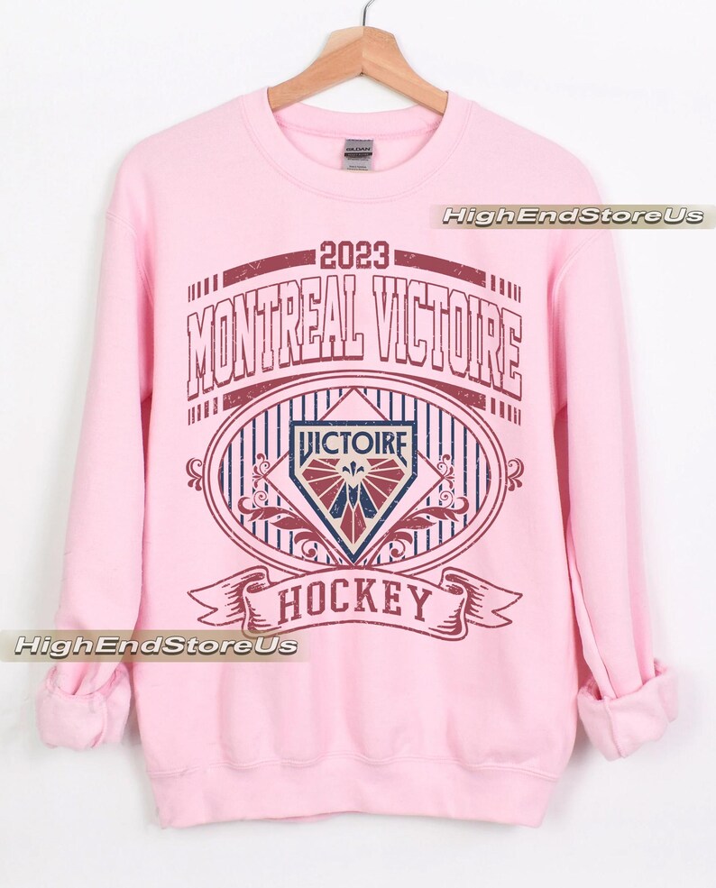 K&ouml;nnte beinhalten: Ein hellrosa Sweatshirt mit einer weinroten und marineblauen Grafik. Die Grafik lautet "2023 MONTREAL VICTOIRE HOCKEY" im Vintage-Stil. Das Sweatshirt ist aus weichem Material und hat lange &Auml;rmel.