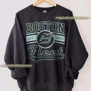 Può includere: Felpa girocollo nera con la scritta "BOSTON Fleet" in un design verde e bianco. La felpa presenta anche la scritta "EST. 2023" e un logo "B" stilizzato. La felpa è appesa a una gruccia di legno.