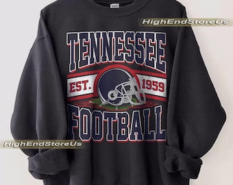 Op de afbeelding: Zwart sweatshirt met de tekst "TENNESSEE FOOTBALL" in witte en rode letters. Een voetbalhelm-afbeelding is gecentreerd boven het woord "FOOTBALL". De tekst "EST. 1959" is ook opgenomen. Het sweatshirt hangt aan een houten hanger.