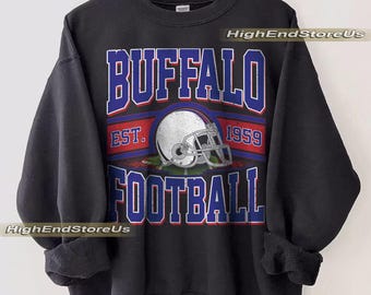 May include: Black sweatshirt with a graphic design featuring the text "BUFFALO FOOTBALL" in blue and white, with a red stripe and a football helmet graphic. The text "EST. 1959" is also included. The sweatshirt is hanging on a wooden hanger.