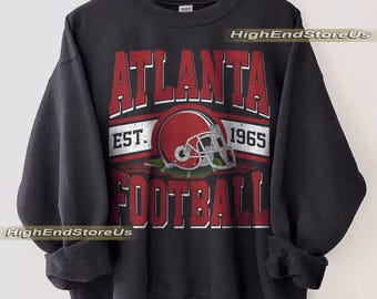 Puede incluir: Sudadera negra con las palabras "ATLANTA FOOTBALL" en letras rojas. Un gráfico de casco de fútbol rojo está centrado debajo de la palabra "ATLANTA". El texto "EST. 1965" está encima del casco. La sudadera está colgada en una percha de madera.