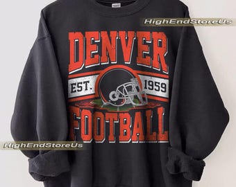 Puede incluir: Sudadera negra con las palabras "DENVER FOOTBALL" en rojo, con un gráfico de casco de fútbol. También se muestra el texto "EST. 1959". La sudadera está colgada en una percha de madera.