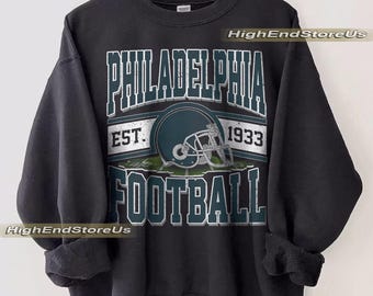 May include: Black sweatshirt with a graphic design featuring the text "PHILADELPHIA FOOTBALL" and a football helmet. The design includes the text "EST. 1933". The sweatshirt is hanging on a wooden hanger.