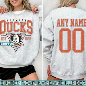 Puede incluir: Sudadera gris con el texto "Anaheim Ducks Est 1967" y un pato de dibujos animados con un casco de hockey. La parte posterior de la sudadera tiene el texto "Any Name 00".