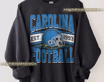 May include: Black sweatshirt featuring a blue and white graphic. The graphic says "CAROLINA FOOTBALL EST. 1993" with a blue football helmet. The text is outlined in white. The sweatshirt is displayed on a wooden hanger.
